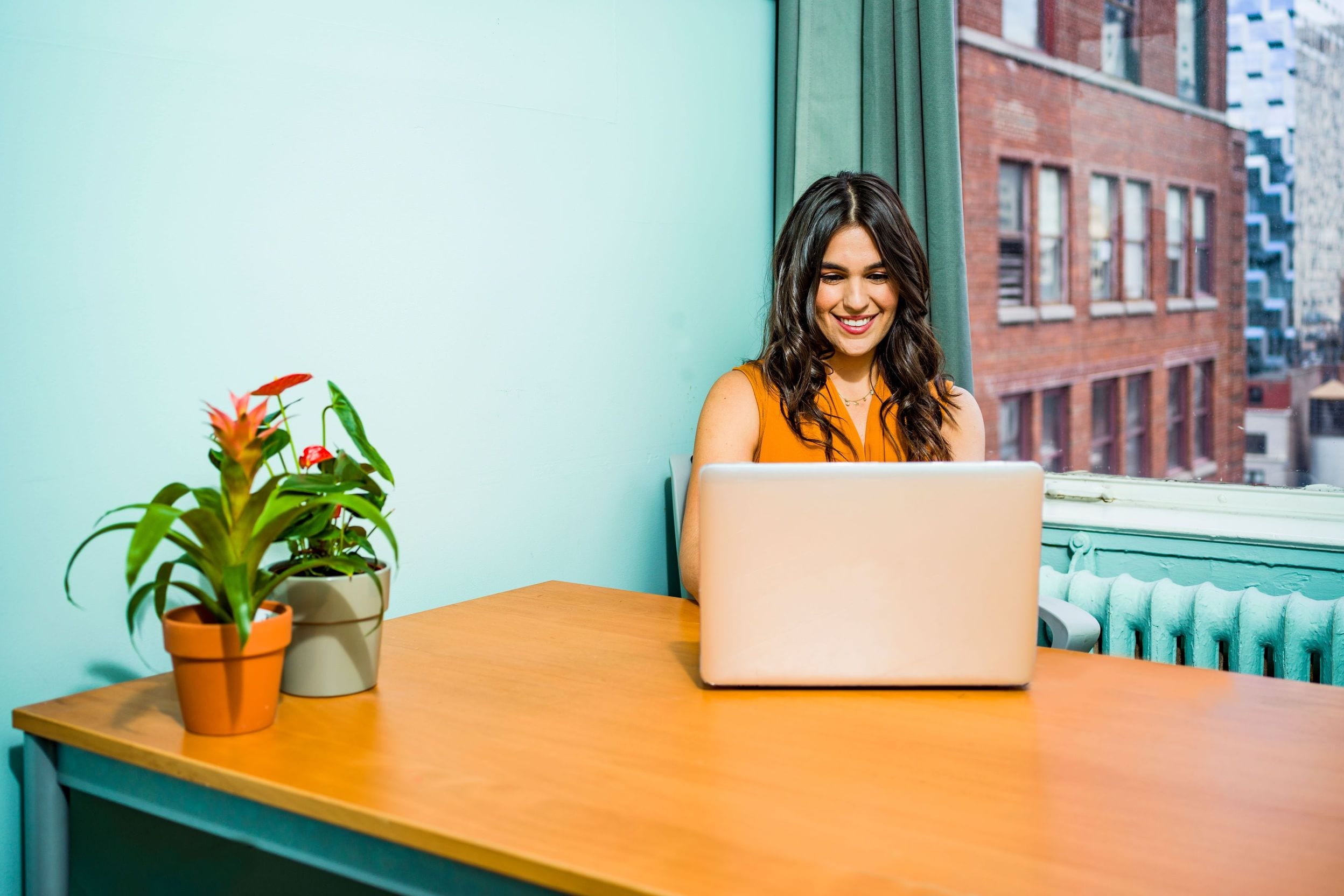 A smiling woman working on her laptop at a wooden desk in a bright office with plants.